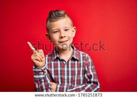 Young little caucasian kid with blue eyes standing wearing elegant shirt over red background with a big smile on face, pointing with hand and finger to the side looking at the camera.