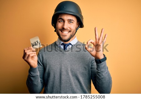 Handsome businessman wearing military helmet holding reminder paper with war message doing ok sign with fingers, excellent symbol