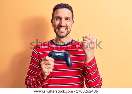 Young handsome gamer man playing video game using gamepad over yellow background screaming proud, celebrating victory and success very excited with raised arm