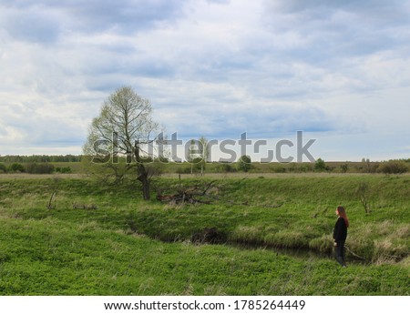 A girl with long red hear admires the scenery with a lonely tree standing on the bank of a small river covered with bright green grass.
