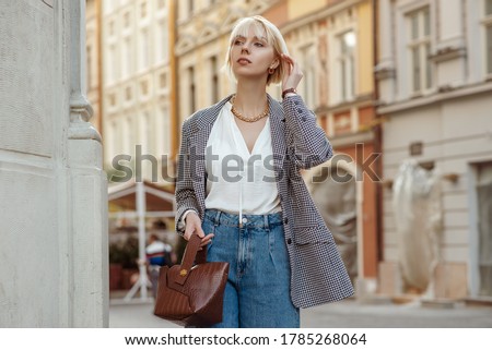 Street fashion photo of elegant  woman wearing trendy white blouse, stylish checkered blazer, high waist jeans, holding brown faux croco leather textured bag. Model walking in street of European city
 Royalty-Free Stock Photo #1785268064
