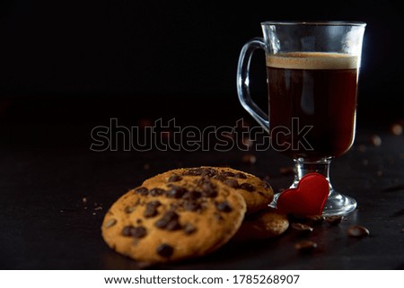 Close up of a glass cup of coffee, chocolate chip cookies and roasted coffee beans on dark background. Concept of ready to eat food, tasty snack. Selective focus, copy space.