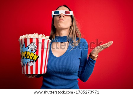 Young blonde woman wearing 3d glasses and eating pack of popcorn watching a movie on cinema crazy and mad shouting and yelling with aggressive expression and arms raised. Frustration concept.