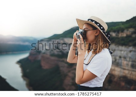young blonde girl in summer hat takes photo on retro camera of panorama mountain landscape walking on trip outdoors, hipster tourist enjoys hobby of photographing nature on holiday