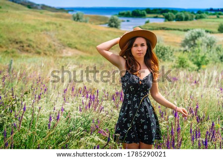 A happy young woman in a dress and hat stands against a background of blooming wildflowers and tall grass in a hilly area