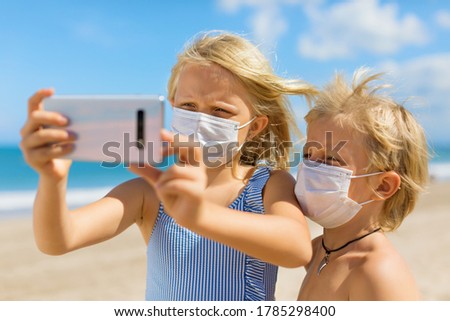 Funny kids taking selfie photo by smartphone on tropical sea beach. New rules to wear cloth face covering mask at public places due coronavirus COVID 19. Family holidays with children, summer travel.