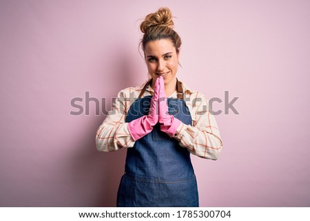 Young beautiful blonde cleaner woman doing housework wearing arpon and gloves praying with hands together asking for forgiveness smiling confident.