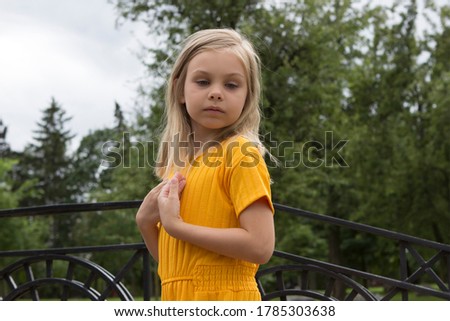 A little girl, a child of six seven years old, blonde with long hair, gray light eyes in yellow bright clothes, lowered her eyes, pressed her hands to her chest, posing like a model. Summer park.