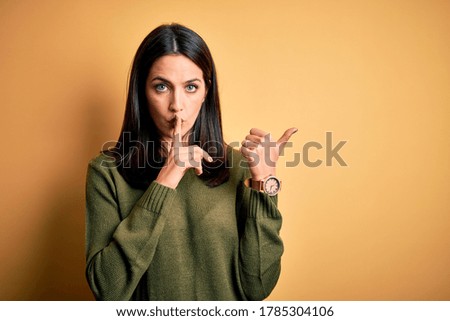 Young brunette woman with blue eyes wearing green casual sweater over yellow background asking to be quiet with finger on lips pointing with hand to the side. Silence and secret concept.