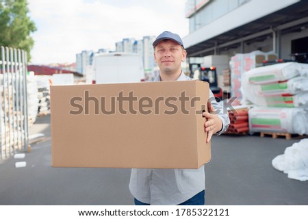 A man in a gray suit in his hands with a cardboard box against the backdrop of a warehouse and a loader. Concept on the topic of postal delivery