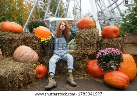 redhead curly Witch holding  orange balloon in hand. Young woman celebrating halloween. Helium party balloon. sit on hay stack. big pumkins lie around. Ferris wheel in park on background.