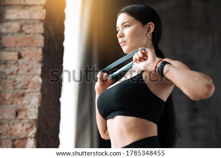 Close-up of a young woman in a black sports tank top doing exercises for the pectoral muscles using an elastic tape expander and looks out the window. Royalty-Free Stock Photo #1785348455