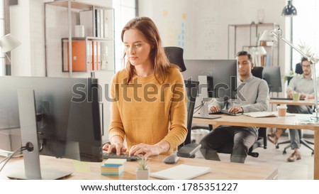 Beautiful and Smart Red Haired Female Specialist Sitting at Her Desk Works on a Desktop Computer. Bright and Modern Open Space Office with Stylish Ergonomic Furniture for Talented Creative People Royalty-Free Stock Photo #1785351227