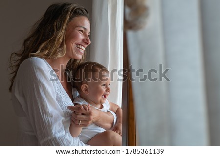 Authentic shot of an young happy smiling neo mother is enjoying time with her newborn baby while looking through a window just woke up in morning in a bedroom.