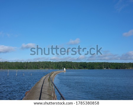 A curved pier with a white marker at the end stretches out into a strait in Sweden. There is blue sky and a distant tree line in the background. The photograph is shot with a medium format camera.