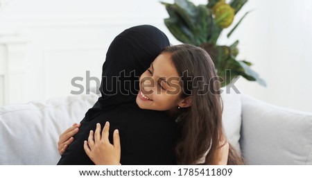 Pretty teen small daughter talking with muslim mother in hijab and embracing. Arab little girl having talk with woman in headscarf on couch at home and hugging. Mom's love hugs. Rear. Back view. Royalty-Free Stock Photo #1785411809