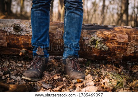 close up photo of human legs in old leather boots and blue jeans standing on the ground with dried leaves