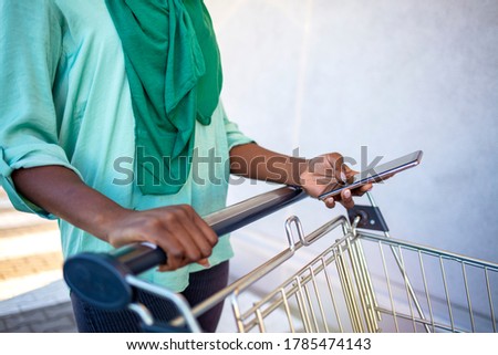 Woman goes grocery with shopping cart. Woman in parking lot pushing a grocery shopping cart and ready to enter the shopping mall. One woman, standing outdoors, using smartphone, shopping cart