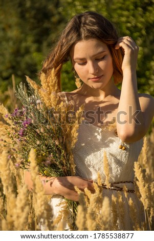 A young Asian girl in a white dress walking on a field with fluffy ears.  A model with a short haircut and a bouquet of wildflowers.