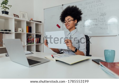 Smiling afro american female teacher showing a blank sheet of paper at web camera of her laptop while teaching English online. Focus on a woman. E-learning. Distance education. English school Royalty-Free Stock Photo #1785553199