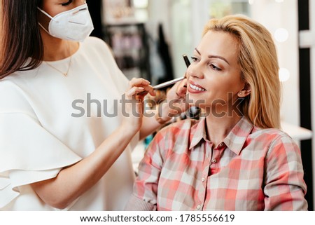 Make up artist with face protective mask applying professional make up of beautiful middle age blonde woman. Coronavirus pandemic lifestyle.