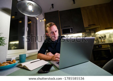 A young businessman sits at a table, holds a smartphone in his hand, uses a laptop and takes notes in his notebook. The guy is working, studying. Online marketing, education, e-learning, e-commerce.