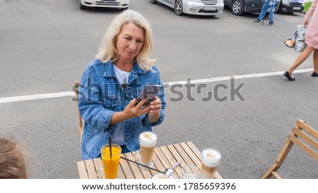 A blonde woman photographs coffee with a cell phone in a street cafe. The woman is wearing a fashionable denim jacket