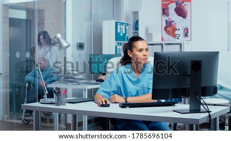 Nurse working on PC in modern private clinic with glass walls where patients are brought in wheelchair from an elevator and other persons are going through hallway. Team of doctors researching Royalty-Free Stock Photo #1785696104