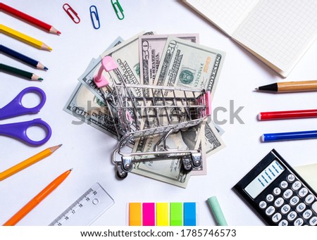 Top view photo of a small shopping cart with dollars in the center and colorful office supplies, isolated on white