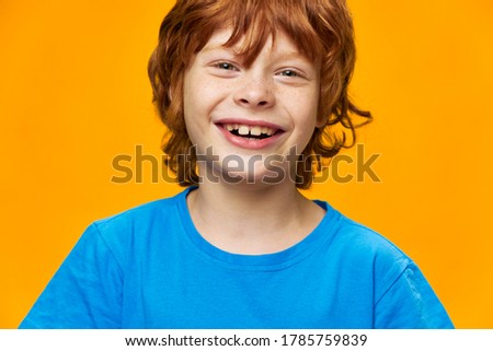 A funny redhead boy smiles into the camera close-up blue t-shirt yellow background 