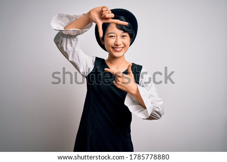Young beautiful asian girl wearing casual dress and hat standing over isolated white background smiling making frame with hands and fingers with happy face. Creativity and photography concept.
