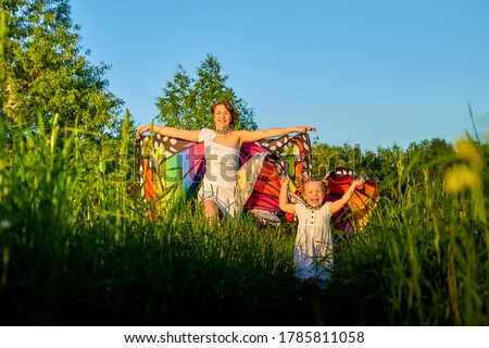 Mother and daughter with bright fabric like butterfly wings play, fly, run, jump in meadow or field with green grass and dandelion flowers. Young woman and a little girl have fun on natural landscape