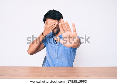 Handsome hispanic man wearing casual clothes sitting on the table covering eyes with hands and doing stop gesture with sad and fear expression. embarrassed and negative concept. 
