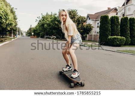 Young woman with long blonde hair, wearing blue denim shorts and white t-shirt, on a long board, on the street.
