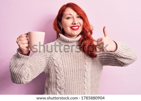 Young beautiful redhead woman drinking cup of coffee over isolated pink background smiling happy and positive, thumb up doing excellent and approval sign