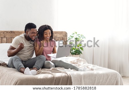 Excited african couple exclaiming happiness, holding laptop and screaming, bedroom interior, free space