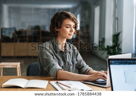 Image of young beautiful focused woman working with laptop while sitting at table in office Royalty-Free Stock Photo #1785884438