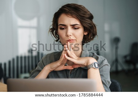 Image of young beautiful brooding woman working with laptop while sitting at table in office Royalty-Free Stock Photo #1785884444