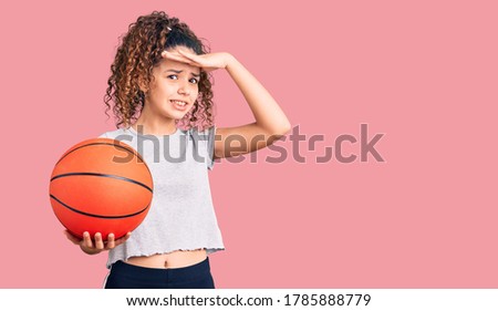 Beautiful kid girl with curly hair holding basketball ball stressed and frustrated with hand on head, surprised and angry face 