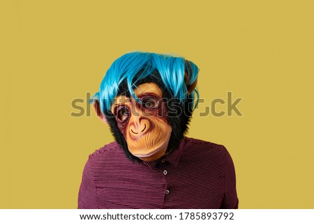 portrait of a man wearing a monkey mask and a blue wig, on a yellow background with some blank space around him