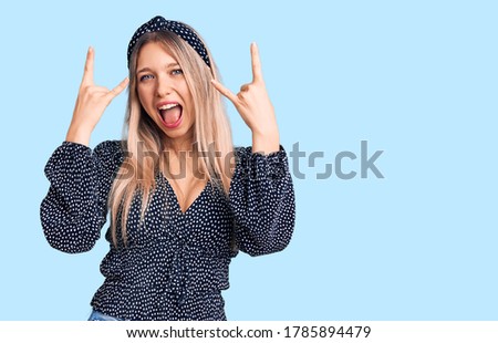 Young beautiful blonde woman wearing casual clothes shouting with crazy expression doing rock symbol with hands up. music star. heavy music concept. 
