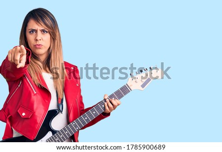 Young beautiful woman playing electric guitar pointing with finger to the camera and to you, confident gesture looking serious 