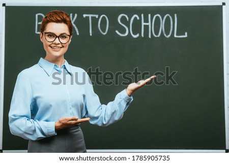 smiling teacher pointing with hands near chalkboard with back to school lettering
