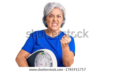 Senior woman with gray hair holding weight machine to balance weight loss annoyed and frustrated shouting with anger, yelling crazy with anger and hand raised  Royalty-Free Stock Photo #1785911717