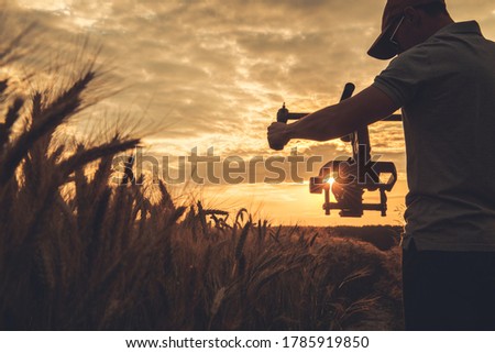 Cinema and Stock Footage Production. Caucasian Video Camera Operator in His 40s with Gimbal Stabilization Taking Steady Scenic Sunset Shot Between Rye Field. Royalty-Free Stock Photo #1785919850