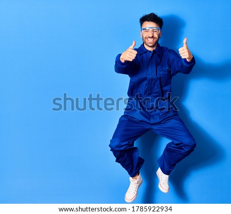 Young handsome hispanic man wearing painter uniform and glasses smiling happy. Jumping with smile on face doing ok sign with thums up over isolated blue background