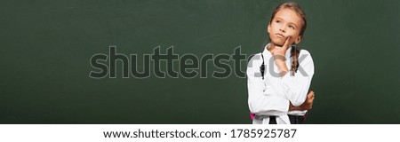 horizontal concept of thoughtful schoolgirl touching face and looking up near chalkboard