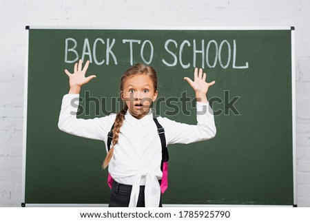 shocked schoolgirl standing with raised hands near chalkboard with back to school inscription