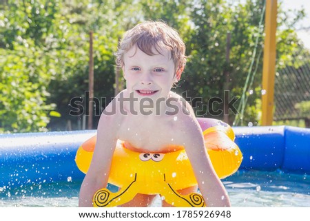 A blond European boy plays in the summer in the pool in nature. A child in a yellow inflatable circle splashes water drops.