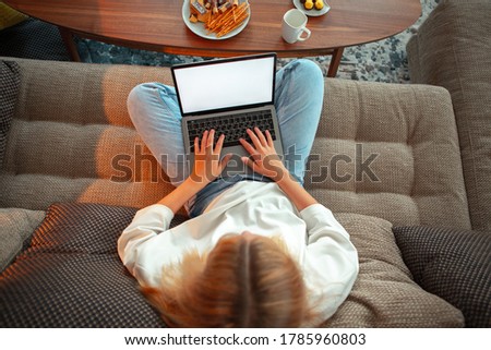 Teen girl sitting on sofa in Lotus position with blank screen laptop watching TV series with sweets and tea, top view